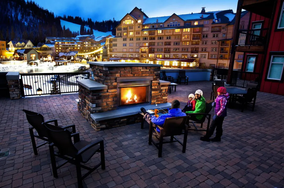 This exterior image features a cozy, inviting seating area centered around a modern outdoor fireplace. Illuminated resort buildings and a snowy landscape provide a festive, luxurious winter ambiance, with the warmth of the fire inviting guests to gather after a day of adventure.