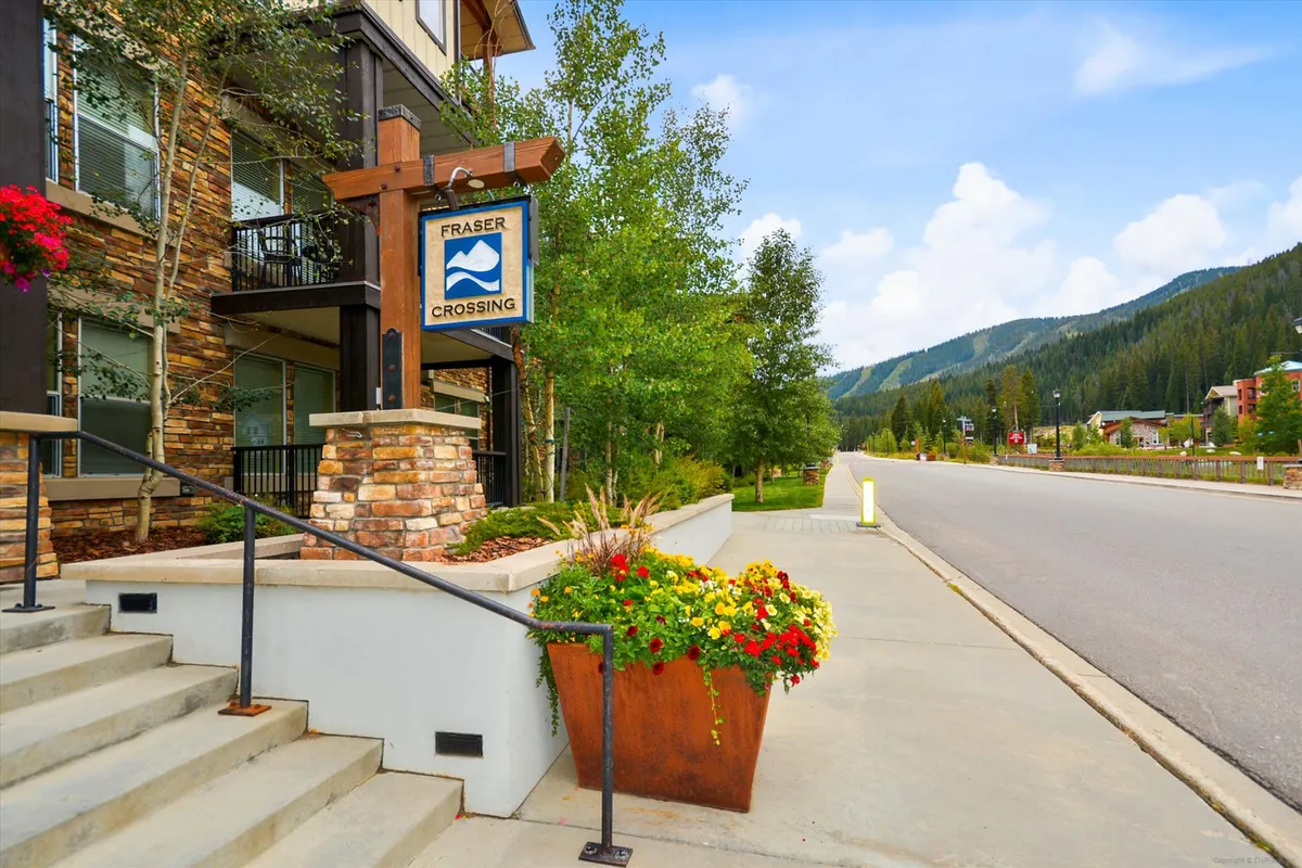 The exterior of Fraser Crossing welcomes guests with lush flower planters, stone steps, and the prominent Fraser Crossing sign surrounded by greenery. People can be seen walking and biking, giving a sense of the vibrant resort village atmosphere and accessibility to outdoor activities.