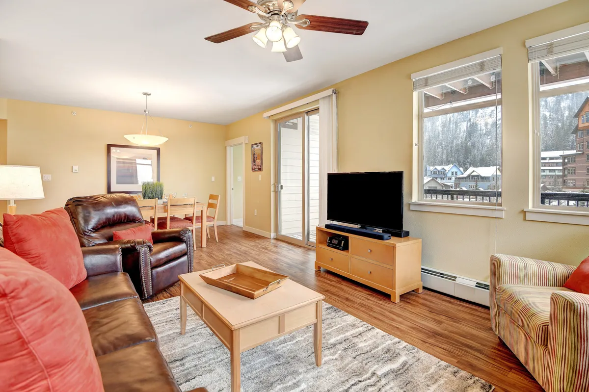 A spacious open-concept living area featuring a comfortable brown leather sofa, vibrant red accent chairs, a wooden coffee table, and a TV stand with a television. Large windows allow natural light to fill the space, while the dining area with chairs is visible in the background. The overall ambiance is cozy and inviting, with designer furnishings enhancing the Rocky Mountain lodge aesthetic.
