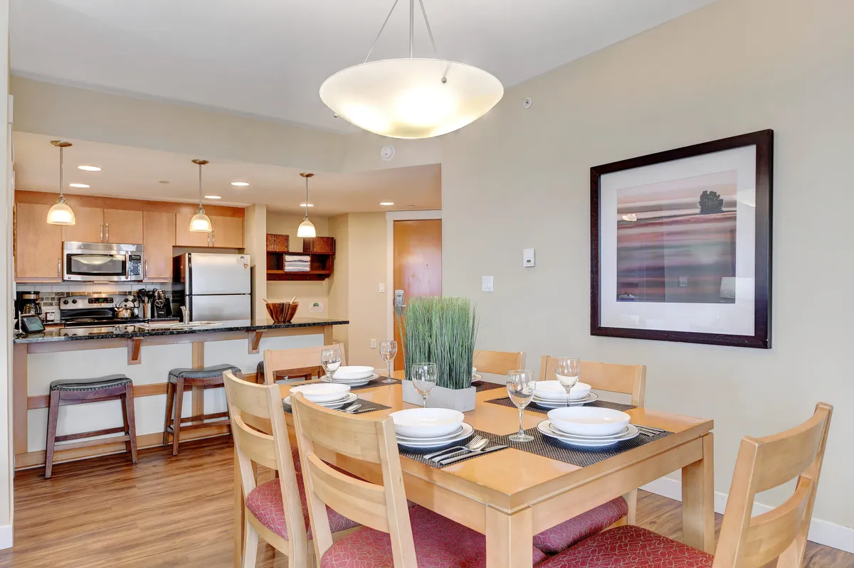 A dining table set with plates and glasses, with the kitchen visible in the background. The space is adorned with warm lighting and tasteful wall art, setting a scene for shared meals and lively conversation.