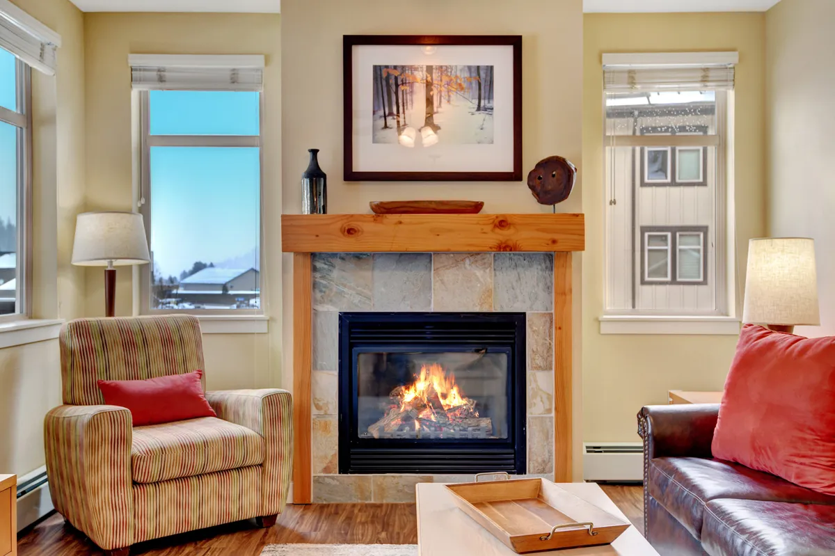 A cozy corner of the living area showcasing a rustic gas fireplace, an inviting armchair, decorative artwork, and sunlit windows that hint at impressive mountain views. The space is arranged for relaxation, with a focus on comfort and warmth by the fire.