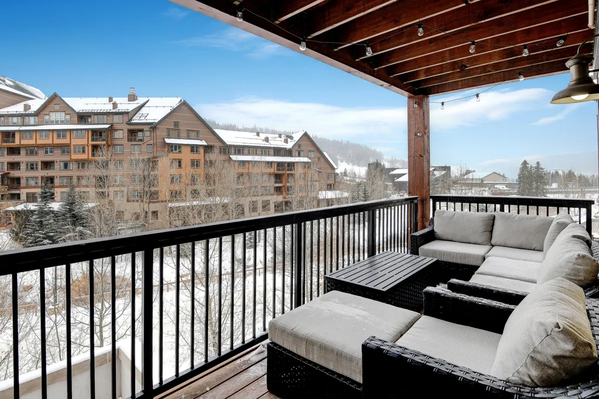 A private covered deck set against a snow-covered landscape, furnished with comfortable patio seating oriented towards a majestic mountain view. The scene is tranquil and inviting, ideal for enjoying morning coffee or evening cocoa while watching the festive village below and patio lights twinkle.
