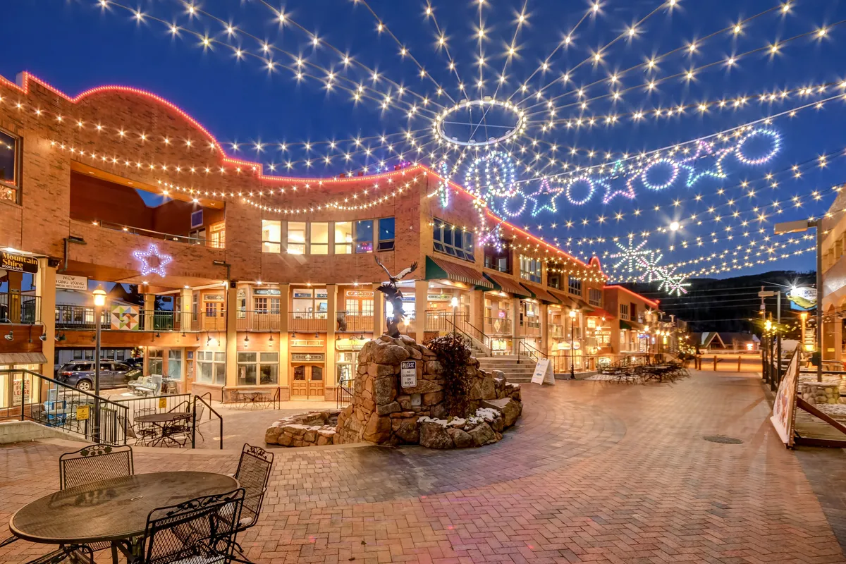 The image shows an exterior village plaza at dusk, decorated with string lights overhead, stone fountains, and vibrant outdoor seating. People are shopping, dining, and enjoying live entertainment, creating a joyful and lively mood that connects directly to the access to resort amenities and the elevated social environment highlighted in the section.