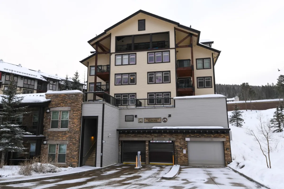 A welcoming view of a multi-story building surrounded by fresh snow, trees, and a parking area. The cozy lights and snow-covered balconies suggest a warm retreat after a day outdoors.