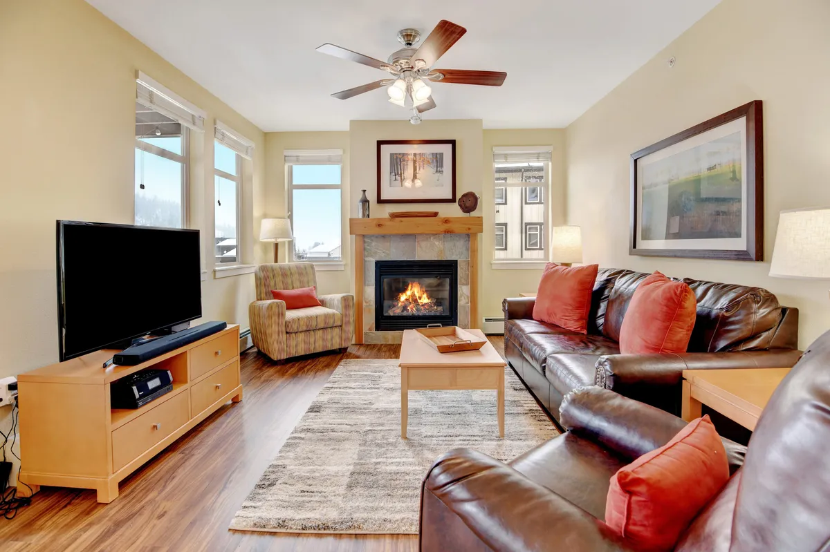 The image captures a cozy, inviting living room centered around a crackling fireplace. Plush sofas and an armchair surround a coffee table, while a large TV is positioned above or near the fireplace. Soft lighting and thoughtfully arranged décor create a warm, relaxing atmosphere perfect for unwinding after a day on the slopes.