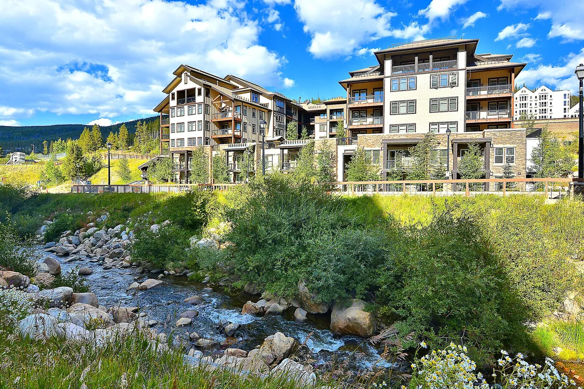 This exterior shot features a modern, multi-story building with private decks overlooking a flowing stream and lush greenery. The peaceful, rejuvenating mood evokes early mornings with coffee on a private deck and evenings spent enjoying nature, aligning with the promise of private, upscale living within a resort context.